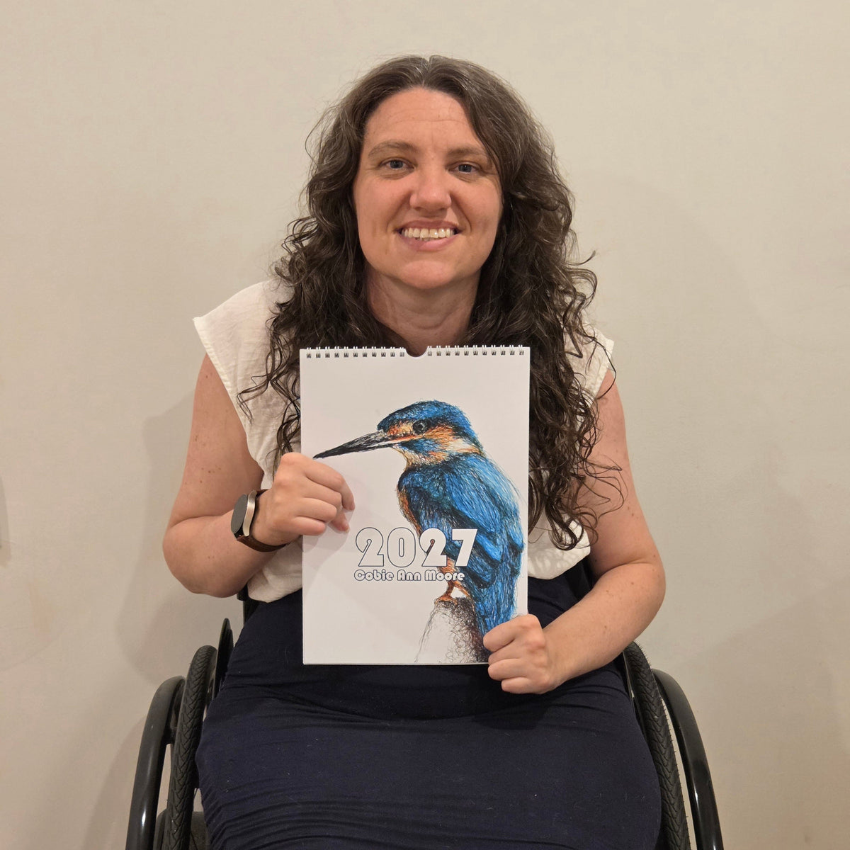 A smiling woman with curly brown hair sits in a wheelchair holding a 2027 spiral-bound calendar featuring a blue kingfisher illustration on the cover, against a plain beige wall.