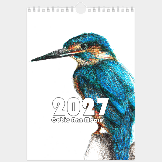 A spiral-bound 2027 wall calendar featuring a detailed colored pencil drawing of a kingfisher on the cover, with the text '2027' and 'Cobie Ann Moore' printed below the bird.