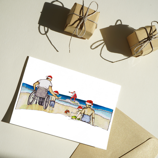 Christmas card with a drawing of a family at the beach watching Santa surfing. The father and one child are on the left side of the card (the father is a wheelchair user). The mother is sitting on the sand with the other child on the right-hand side of the card.